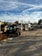 An outdoor industrial worksite is shown with a forklift in the foreground. The area contains piles of wooden planks and other materials. In the background, there are structures including storage containers and various equipment.