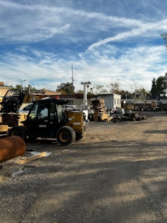 An outdoor industrial worksite is shown with a forklift in the foreground. The area contains piles of wooden planks and other materials. In the background, there are structures including storage containers and various equipment.