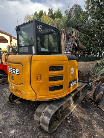 A yellow John Deere mini excavator is parked outdoors. It has a compact tracked undercarriage and a black cab with windows, surrounded by lush green palm trees and foliage on all sides.