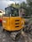 A yellow John Deere mini excavator is parked outdoors. It has a compact tracked undercarriage and a black cab with windows, surrounded by lush green palm trees and foliage on all sides.