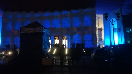 A building facade is illuminated with blue and white lights, creating an artistic effect. Silhouettes of people and vehicles are visible in the foreground, with more people near the entrance. The lighting gives the scene a dramatic and vivid appearance.