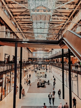 A bustling indoor shopping area featuring a high open ceiling with exposed steel beams. The scene includes multiple levels with walkways, numerous shops with bright signage, and a variety of people walking and browsing. The central area of the floor shows an informational or vendor booth and a parked vehicle.