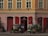 A charming storefront with red shutters and an array of potted plants and flowers displayed on racks and the pavement. The building features a muted orange facade with several windows, and there are light fixtures and decorative elements above the entrance.