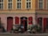 A charming storefront with red shutters and an array of potted plants and flowers displayed on racks and the pavement. The building features a muted orange facade with several windows, and there are light fixtures and decorative elements above the entrance.