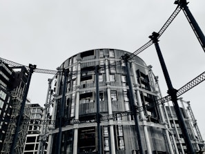 A circular modern building with intricate metal framework standing among other contemporary architectural structures. The steel beams provide an industrial aesthetic amidst a gray, overcast sky.