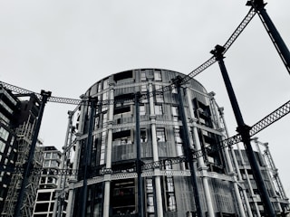 A circular modern building with intricate metal framework standing among other contemporary architectural structures. The steel beams provide an industrial aesthetic amidst a gray, overcast sky.