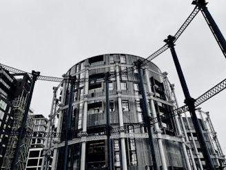 A circular modern building with intricate metal framework standing among other contemporary architectural structures. The steel beams provide an industrial aesthetic amidst a gray, overcast sky.