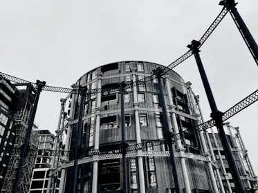 A circular modern building with intricate metal framework standing among other contemporary architectural structures. The steel beams provide an industrial aesthetic amidst a gray, overcast sky.