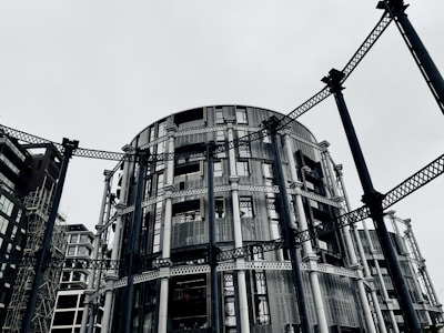 A circular modern building with intricate metal framework standing among other contemporary architectural structures. The steel beams provide an industrial aesthetic amidst a gray, overcast sky.