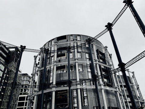 A circular modern building with intricate metal framework standing among other contemporary architectural structures. The steel beams provide an industrial aesthetic amidst a gray, overcast sky.