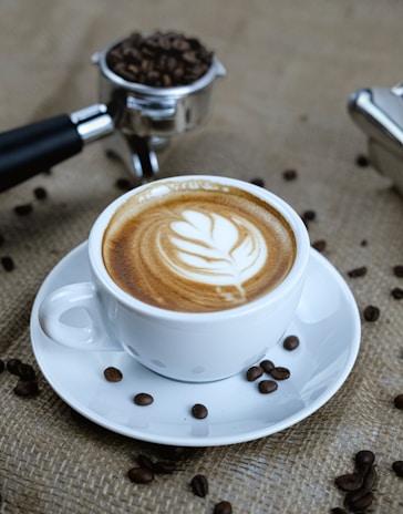 A white cup containing a latte with intricate leaf art on the frothy surface sits on a saucer. Scattered coffee beans surround the cup, and a metal portafilter filled with more coffee beans is visible in the background on a textured burlap surface.