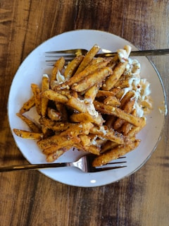 A plate of seasoned French fries covered with spices and creamy sauce is placed on a wooden table. The fries are golden brown and appear crispy, with some red chili flakes scattered on top. Two forks are placed on the plate, suggesting it's meant for sharing.