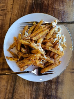 A plate of seasoned French fries covered with spices and creamy sauce is placed on a wooden table. The fries are golden brown and appear crispy, with some red chili flakes scattered on top. Two forks are placed on the plate, suggesting it's meant for sharing.