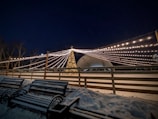 A nighttime scene features a large, lit Christmas tree beneath a canopy of string lights that stretch outwards, creating a festive atmosphere. Snow covers the ground and benches are lined up along a wooden fence, adding to the wintry setting.