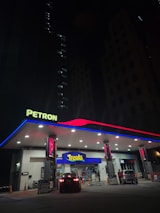 A well-lit Petron gas station at night features illuminated signage and a canopy with red and blue accents. Two cars are parked near the pumps with one heading towards the convenience store labeled 'Treats'. The scene is framed by dark, tall buildings in the background, with only a few lights visible in the windows.