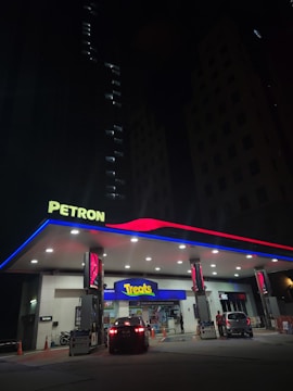 A well-lit Petron gas station at night features illuminated signage and a canopy with red and blue accents. Two cars are parked near the pumps with one heading towards the convenience store labeled 'Treats'. The scene is framed by dark, tall buildings in the background, with only a few lights visible in the windows.