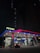 A well-lit Petron gas station at night features illuminated signage and a canopy with red and blue accents. Two cars are parked near the pumps with one heading towards the convenience store labeled 'Treats'. The scene is framed by dark, tall buildings in the background, with only a few lights visible in the windows.