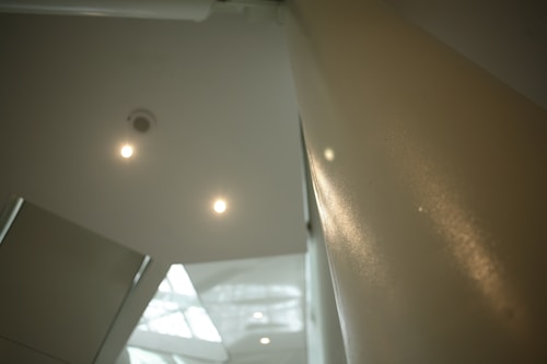 The image features an abstract architectural interior, including parts of a ceiling with small lights, a beige wall, and a glimpse of an angular structure with large windows that allow natural light to illuminate the area.