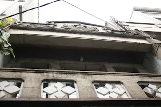 An upward view of a building facade featuring decorative stonework and geometric window designs. The structure appears aged with visible wear on the surfaces. Greenery can be seen on the left side, adding a touch of nature to the urban setting.