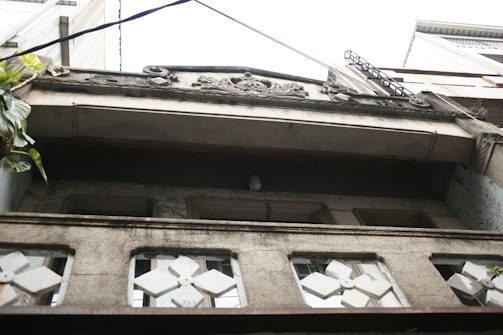 An upward view of a building facade featuring decorative stonework and geometric window designs. The structure appears aged with visible wear on the surfaces. Greenery can be seen on the left side, adding a touch of nature to the urban setting.