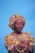 A person wearing a vibrant yellow and brown patterned traditional outfit, including a headwrap and matching dress. They are holding a brown handbag with gold chain details. The background is a solid light blue, and the person is accessorized with gold jewelry, including earrings and a necklace.