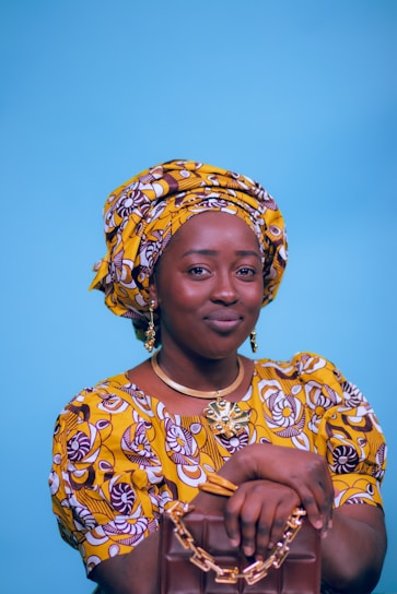 A person wearing a vibrant yellow and brown patterned traditional outfit, including a headwrap and matching dress. They are holding a brown handbag with gold chain details. The background is a solid light blue, and the person is accessorized with gold jewelry, including earrings and a necklace.