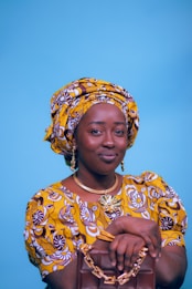 A person wearing a vibrant yellow and brown patterned traditional outfit, including a headwrap and matching dress. They are holding a brown handbag with gold chain details. The background is a solid light blue, and the person is accessorized with gold jewelry, including earrings and a necklace.