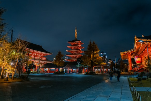 A nighttime scene features a beautifully illuminated traditional Japanese temple with multiple buildings and a pagoda. The structures are adorned with red accents and warm lighting, highlighting intricate architectural details. The dark sky contrasts with the ambient lighting, and a few people can be seen walking along the stone pathway, adding a sense of scale and life to the serene environment.
