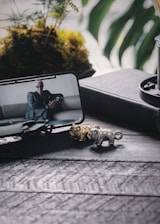 A smartphone displays a suited person sitting on a couch, positioned on a dark textured surface. In front of the phone, small metallic animal figurines in gold and silver are visible. A pot with a potted plant containing lush greenery and moss is in the background, adding a natural element.