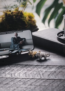 A smartphone displays a suited person sitting on a couch, positioned on a dark textured surface. In front of the phone, small metallic animal figurines in gold and silver are visible. A pot with a potted plant containing lush greenery and moss is in the background, adding a natural element.
