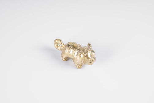 A small, golden animal figurine with detailed fur texture placed on a plain white surface. The figure appears to have a curled tail and upright ears.