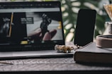 A laptop displaying a website is open on a wooden table. Next to it is a smartphone with a dark screen. In front of the laptop, there is a small gold and silver figurine resembling a cat. Beside these items, a large book lies closed. Green leaves in the background add a touch of nature.