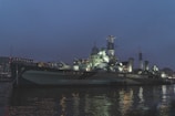 An illuminated naval warship is docked at a harbor during twilight. The ship's structure includes multiple decks, large guns, antennas, and radar equipment. The surrounding environment is dimly lit, with reflections of the ship's lights glimmering on the water's surface. In the background, there are modern buildings with lit windows.