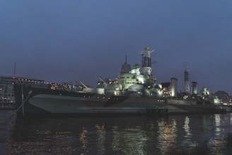 An illuminated naval warship is docked at a harbor during twilight. The ship's structure includes multiple decks, large guns, antennas, and radar equipment. The surrounding environment is dimly lit, with reflections of the ship's lights glimmering on the water's surface. In the background, there are modern buildings with lit windows.