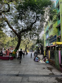A bustling urban street with trees lining the sidewalk and people walking. A few individuals appear to be sitting on the ground next to a building selling items. The building is painted in vibrant green and yellow. The scene is illuminated by daylight, and there is a sense of daily life occurring in the city.