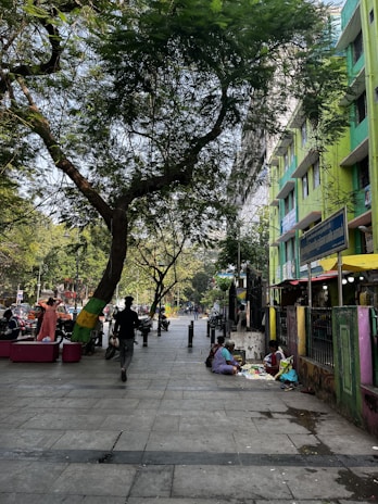 A bustling urban street with trees lining the sidewalk and people walking. A few individuals appear to be sitting on the ground next to a building selling items. The building is painted in vibrant green and yellow. The scene is illuminated by daylight, and there is a sense of daily life occurring in the city.