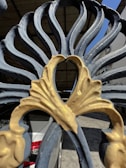 An ornate metal gate with an intricate leaf design featuring blue and gold colors. The pattern has a heart-like center and allows partial visibility of a white car parked in a garage and surrounding structures.