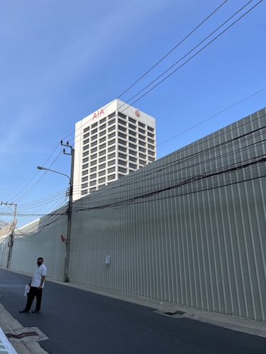 A tall white building with the letters 'AIA' on top stands against a clear blue sky. The building is surrounded by metal fences. A person wearing a white shirt and black pants walks along a quiet street, which is lined with overhead utility wires.