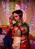 A person is dressed in traditional attire, adorned with elaborate jewelry and a decorative headpiece. They are holding two betel leaves in front of their face, elegantly detailed with intricate designs. The background features colorful floral and leaf patterns, contributing to a festive atmosphere.