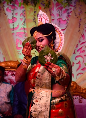A person is dressed in traditional attire, adorned with elaborate jewelry and a decorative headpiece. They are holding two betel leaves in front of their face, elegantly detailed with intricate designs. The background features colorful floral and leaf patterns, contributing to a festive atmosphere.