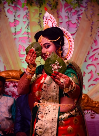 A person is dressed in traditional attire, adorned with elaborate jewelry and a decorative headpiece. They are holding two betel leaves in front of their face, elegantly detailed with intricate designs. The background features colorful floral and leaf patterns, contributing to a festive atmosphere.