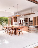 A modern kitchen and dining area with an open layout, featuring wooden cabinetry and furniture. The space includes a long dining table with matching chairs, pendant lights above, and a large kitchen island. Natural light streams in from sliding glass doors that open to a garden with bamboo plants.
