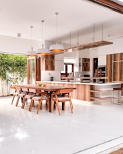 A modern kitchen and dining area with an open layout, featuring wooden cabinetry and furniture. The space includes a long dining table with matching chairs, pendant lights above, and a large kitchen island. Natural light streams in from sliding glass doors that open to a garden with bamboo plants.
