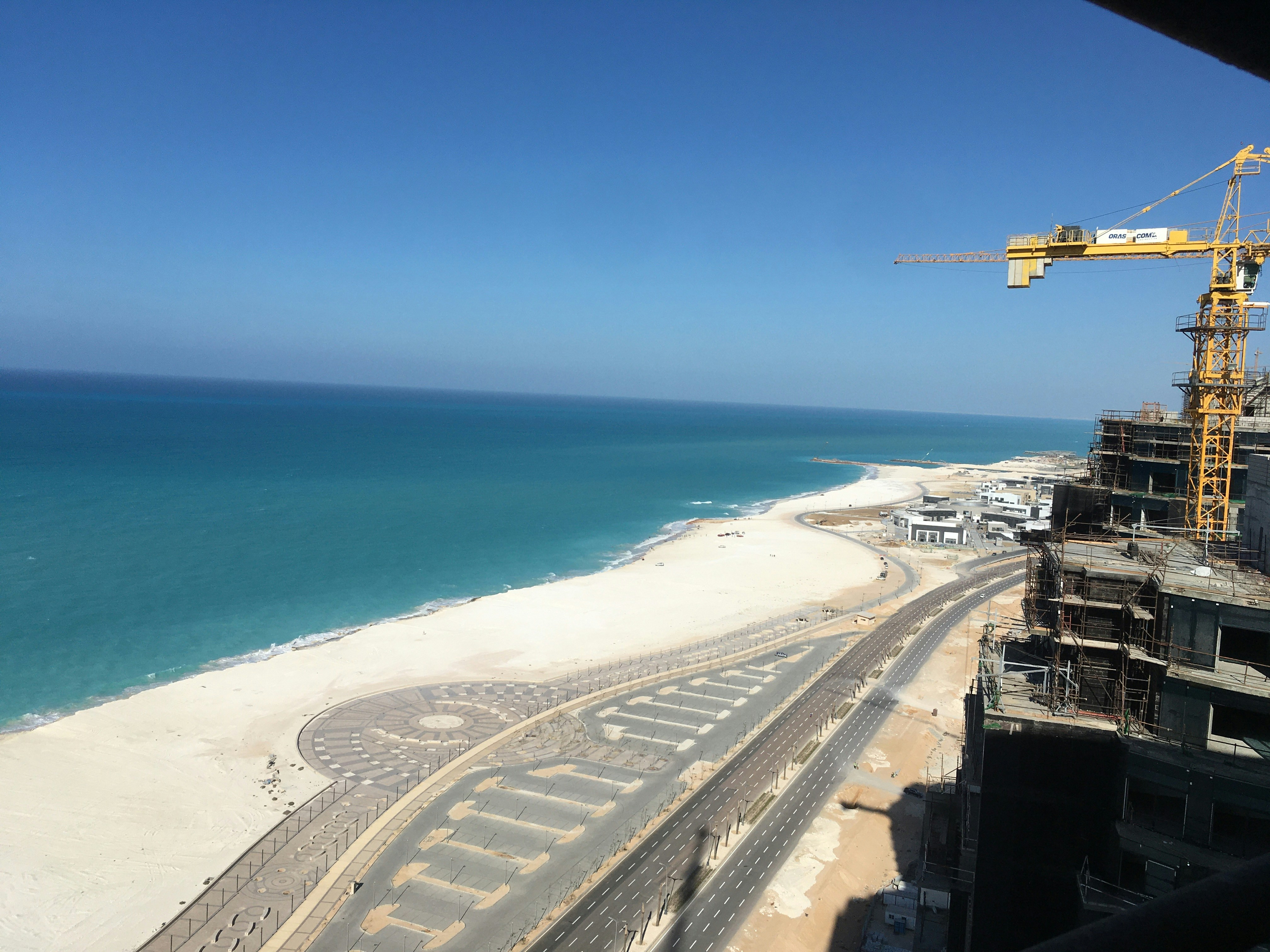 A coastal construction site with a view of the sea. A large yellow crane towers over partially constructed buildings. The area features a newly paved road running parallel to the beach, with clear weather and an expansive blue sky.