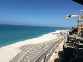 A coastal construction site with a view of the sea. A large yellow crane towers over partially constructed buildings. The area features a newly paved road running parallel to the beach, with clear weather and an expansive blue sky.