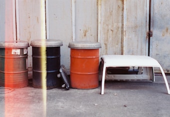 Three oil drums with metal lids in green, black, and orange are lined up against a perforated metal backdrop. Beside them is a white plastic table resting sideways. The ground is concrete, and there is a vertical strip of red and yellow light on the left side, suggesting light leakage on the photograph.
