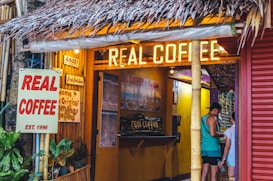A cozy coffee shop with a tropical vibe features a thatched roof and warm yellow lighting. The signage includes bold lettering stating 'REAL COFFEE' and additional decor promoting various items like fresh fruit juice. Two people are visible at the counter, surrounded by vibrant plants and bamboo structures.