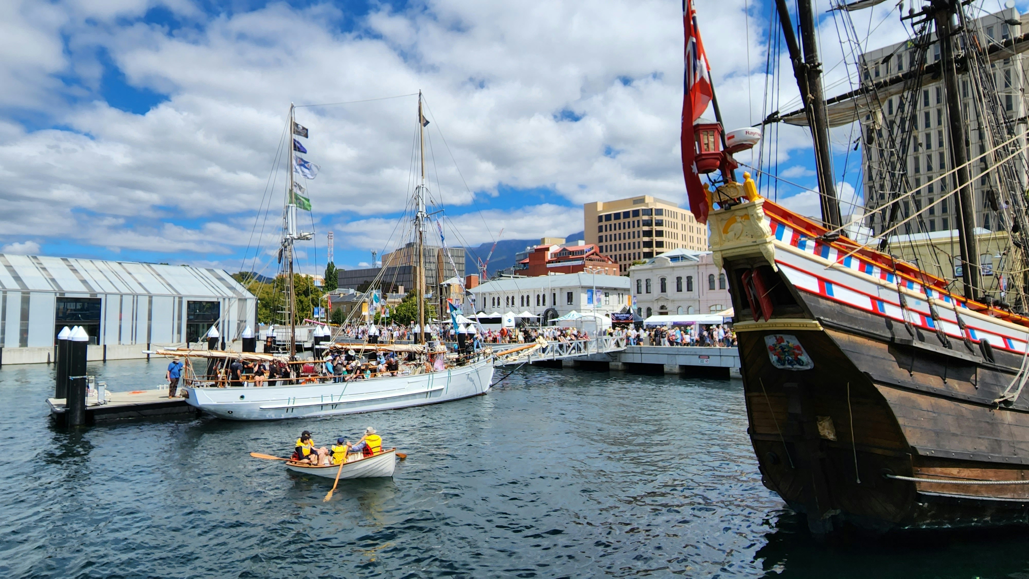 People gather at a busy harbor with historic ships.