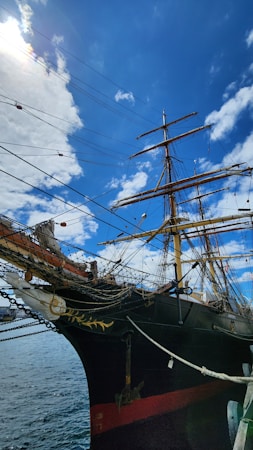 A classic wooden sailing ship with multiple masts and intricate rigging set against a bright blue sky with some scattered clouds. The ship's hull is black with detailed carvings near the bow, and an anchor hangs from the side. The sunlight is creating a lens flare effect in the sky.