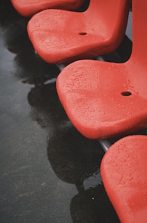 Red plastic chairs with droplets of water on their surface, indicating recent rain. The chairs are arranged in a row and their reflection is visible on the wet ground below.